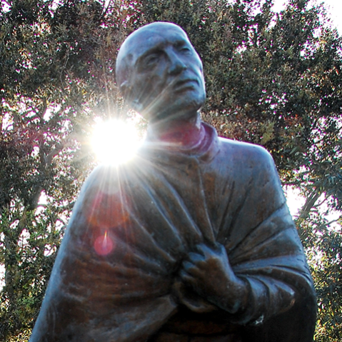 A statue of St. Ignatius from the waist up, with bushes in the background, and the sun peaking out through the leaves above his right shoulder.